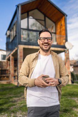 40 yaşında yetişkin bir adam elinde bir fincan çay ya da kahveyle modern evinin önünde güvenle duruyor hayatından memnun ve mutlu görünüyor ve evinin fotokopi alanından gurur duyuyor.
