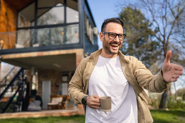 一位40岁的成年人自信地站在他的现代住宅前 微笑着品茶或喝咖啡 他看上去对自己的生活感到满意和快乐 并为自己的房子空间感到自豪 — 图库照片