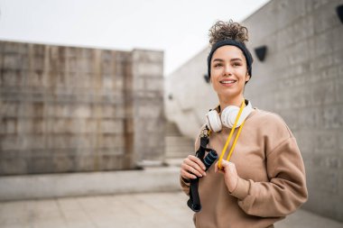 Kafkasyalı genç bir kadının portresi modern beton duvarın önünde duruyor. Kulaklıkları ve lastik direnç bantları ile kendine güvenen kulaklıkları var.