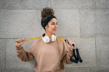 Kafkasyalı genç bir kadının portresi modern beton duvarın önünde duruyor. Kulaklıkları ve lastik direnç bantları ile kendine güvenen kulaklıkları var.