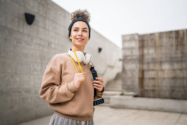 Portrait of one young adult caucasian woman stand outdoor in front of modern concrete wall with headphones confident with rubber resistance band tubes copy space