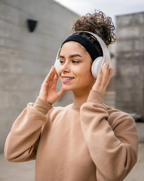 一人の若い大人の白人女性の肖像画は 彼女の頭の上にヘッドフォンで現代のコンクリート壁の前に屋外に立つ自信を持って音楽を聞く — ストック写真