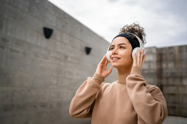 一人の若い大人の白人女性の肖像画は 彼女の頭の上にヘッドフォンで現代のコンクリート壁の前に屋外に立つ自信を持って音楽を聞く — ストック写真