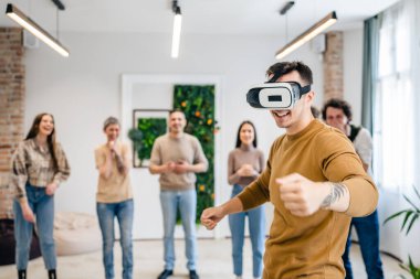 Yetişkin bir beyaz erkek, bir grup erkek ve kadın arkadaşın önünde sanal gerçeklik cihazının keyfini çıkarıyor. Takım oluşturma semineri sırasında birlikte eğleniyorlar. Gerçek insanlar parlak filtreler.