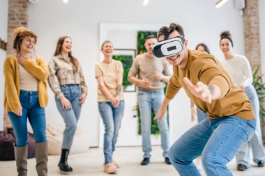 Yetişkin bir beyaz erkek, bir grup erkek ve kadın arkadaşın önünde sanal gerçeklik cihazının keyfini çıkarıyor. Takım oluşturma semineri sırasında birlikte eğleniyorlar. Gerçek insanlar parlak filtreler.