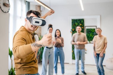 Yetişkin bir beyaz erkek, bir grup erkek ve kadın arkadaşın önünde sanal gerçeklik cihazının keyfini çıkarıyor. Takım oluşturma semineri sırasında birlikte eğleniyorlar. Gerçek insanlar parlak filtreler.