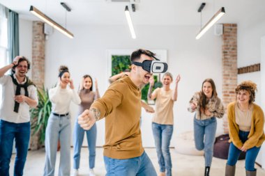 Yetişkin bir beyaz erkek, bir grup erkek ve kadın arkadaşın önünde sanal gerçeklik cihazının keyfini çıkarıyor. Takım oluşturma semineri sırasında birlikte eğleniyorlar. Gerçek insanlar parlak filtreler.