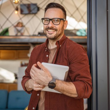 Yetişkin bir beyaz erkek balkonda dikiliyor. Dijital tablet mutlu bir gülümseme tutuyor. Kapının yanında duruyor.