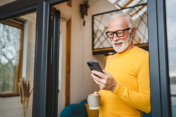 stock image One senior man with a beard stand on the balcony hold a mobile phone and cup of coffee happy smile daily morning routine real person copy space read or use smartphone internet browse social network