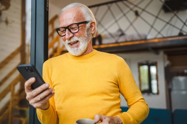 stock image One senior man with a beard stand on the balcony hold a mobile phone and cup of coffee happy smile daily morning routine real person copy space read or use smartphone internet browse social network