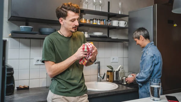 大人の白人男性は台所に立っているカップと布を保持しながら 彼の母親は接続と結合の特別な瞬間を作成する会話に従事する料理を世話一緒に仕事 — ストック写真