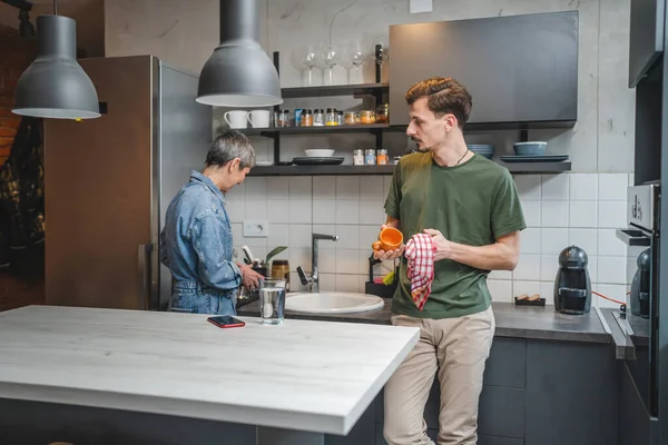 大人の白人男性は台所に立っているカップと布を保持しながら 彼の母親は接続と結合の特別な瞬間を作成する会話に従事する料理を世話一緒に仕事 — ストック写真