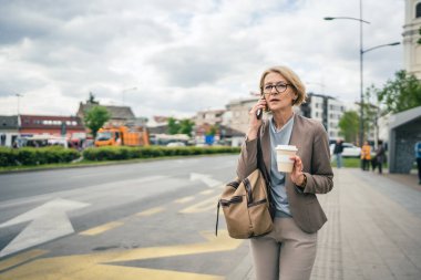 Kafkasyalı, gözlüklü, yetişkin bir kadın otobüs durağındaki durakta duruyor. Akıllı telefon kullanıyor, telefonla konuşuyor ve kötü haberler alıyor.