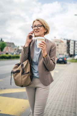 Kafkasyalı, gözlüklü, yetişkin bir kadın otobüs durağında duruyor. Akıllı telefon kullanıyor, telefonla konuşuyor. Gerçek bir insanın işe geç gelmesini bekliyor.