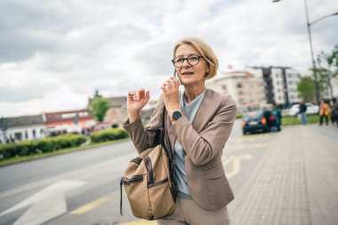 Kafkasyalı, gözlüklü, yetişkin bir kadın otobüs durağında duruyor. Akıllı telefon kullanıyor, telefonla konuşuyor. Gerçek bir insanın işe geç gelmesini bekliyor.