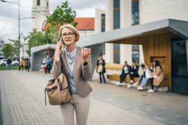 Kafkasyalı, gözlüklü, yetişkin bir kadın otobüs durağında duruyor. Akıllı telefon kullanıyor, telefonla konuşuyor. Gerçek bir insanın işe geç gelmesini bekliyor.