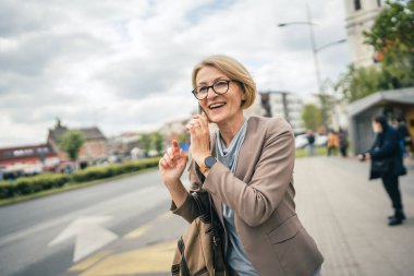Kafkasyalı, gözlüklü, yetişkin bir kadın otobüs durağında duruyor. Akıllı telefon kullanıyor, telefonla konuşuyor. Gerçek bir insanın işe geç gelmesini bekliyor.