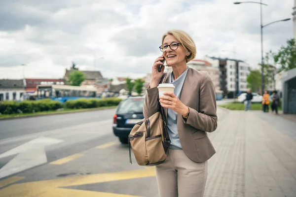 1人の女性の原因アジアの成熟したブロンド女性で眼鏡をかけた女性は バスの街の停留所で一日で停止しますスマートフォンの携帯電話を使用して通話をしながら ドライブを待っています実際の人のコピースペース仕事のために遅い — ストック写真