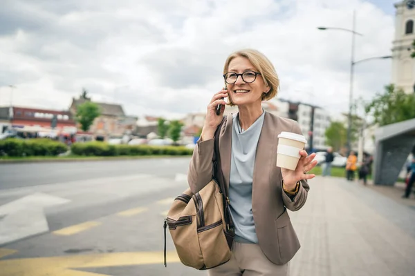 一个女人带着眼镜站在公共汽车站台上 用智能手机打电话 一边等司机 一边等着司机把车位抄送去上班 她是个成熟的金发女人 — 图库照片