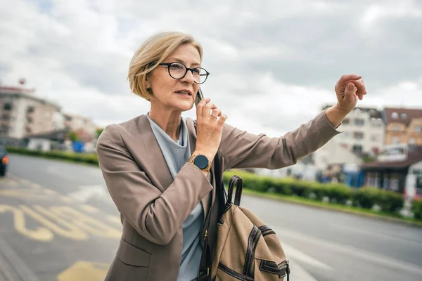 1人の女性の原因アジアの成熟したブロンド女性で眼鏡をかけた女性は バスの街の停留所で一日で停止しますスマートフォンの携帯電話を使用して通話をしながら ドライブを待っています実際の人のコピースペース仕事のために遅い — ストック写真