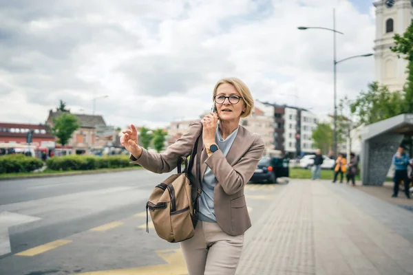 一个女人带着眼镜站在公共汽车站台上 用智能手机打电话 一边等司机 一边等着司机把车位抄送去上班 她是个成熟的金发女人 — 图库照片
