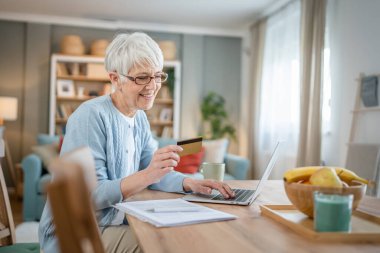 Yetişkin bir yaşlı kadın büyükanne evde oturup online alışveriş siteleri için kredi kartı ya da banka kartı kullanıyor. İndirimli indirimli ürünler alıyor. Gerçek insanlar bilgisayar kopyalıyor.