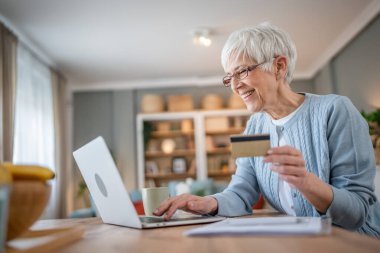 Yetişkin bir yaşlı kadın büyükanne evde oturup online alışveriş siteleri için kredi kartı ya da banka kartı kullanıyor. İndirimli indirimli ürünler alıyor. Gerçek insanlar bilgisayar kopyalıyor.