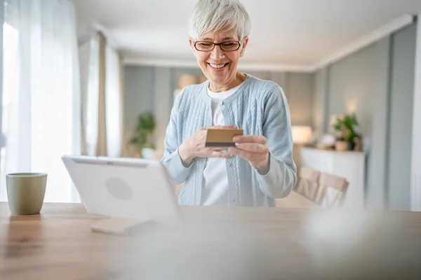 ある成熟した年配の女性の祖母はオンラインショッピングのための家の使用のクレジットカードかデビットカードに坐る実際の人々がスペースをコピーするものを買うインターネット店 — ストック写真
