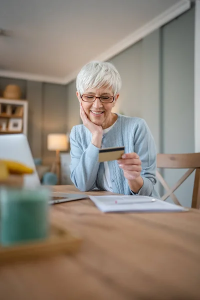 ある成熟した年配の女性の祖母はオンラインショッピングのための家の使用クレジットカードかデビットカードに坐るインターネットの店は割引販売の事を買う実際の人々はノートパソコンのコンピュータを使用するスペースをコピーする — ストック写真