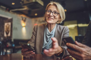 Gözlük takan şık, olgun, sarışın bir kadın kafeteryada rahatça oturup akıllı telefonuyla internete mesaj atıyor ve olgun bireylerin dijital etkileşimlerini inceliyor.