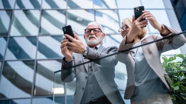 Takım elbise giymiş kıdemli bir adam ve iş ortağı olarak olgun sarışın bir kadın karı koca ya da iş arkadaşı olarak konuşuyorlar akıllı telefonlar ellerinde internetteki internet fotoğraflarına bakıyorlar.