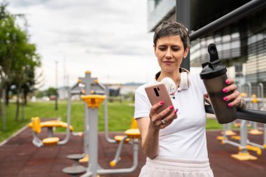 Beyaz bir kadın açık hava antrenmanı sırasında fren yapıyor. Açık hava spor salonunda barlarda dinleniyor. İçme suyu ya da takviye su ile dinleniyor.