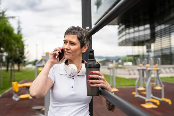 Beyaz bir kadın açık hava antrenmanı sırasında fren yapıyor. Açık hava spor salonunda barlarda dinleniyor. İçme suyu ya da takviye su ile dinleniyor.