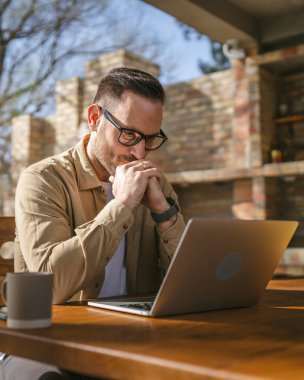 Kafkasyalı modern bir erkek balkon terasında tek başına oturuyor tişört giyiyor dizüstü bilgisayarda çalışıyor uzaktan gülüyor kendine güvenen başarı kavramını taklit ediyor.