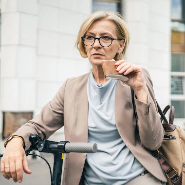 Ciddi bir yetişkin beyaz kadın iş kadını dışarıda elinde elektrikli scooter şişesiyle dikiliyor. Depresyondaki sert yaşam konseptini içiyor. Alkol bağımlılığı alkolizmi kopyalıyor.