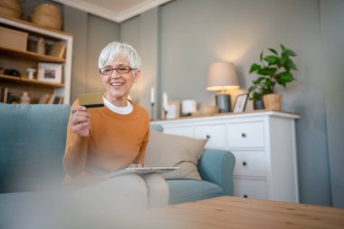 Yetişkin bir yaşlı kadın büyükanne evde oturup online alışveriş siteleri için kredi kartı ya da banka kartı kullanıyor. İndirimli indirimli ürünler alıyor. Gerçek insanlar dijital tablet kullanıyor.