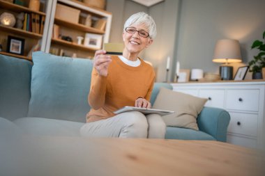 Yetişkin bir yaşlı kadın büyükanne evde oturup online alışveriş siteleri için kredi kartı ya da banka kartı kullanıyor. İndirimli indirimli ürünler alıyor. Gerçek insanlar dijital tablet kullanıyor.