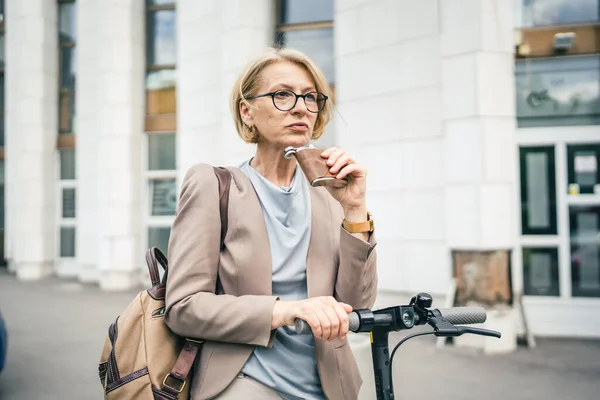 One woman serious mature caucasian female businesswoman stand outdoor with electric kick scooter hold flask drinking desperate depressed hard life concept alcohol abuse alcoholism copy space