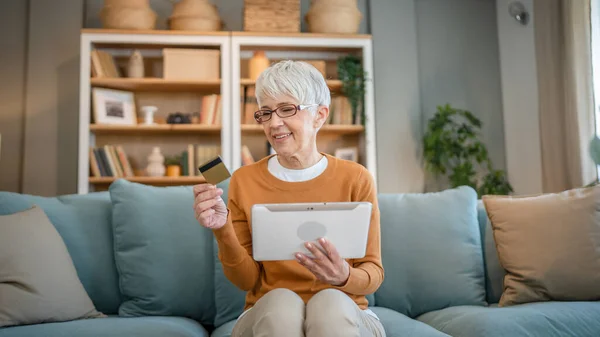 ある成熟した年配の女性の祖母はオンラインショッピングのための家の使用クレジットカードかデビットカードに坐るインターネットの店は割引販売の事を買う実際の人々のコピースペースはデジタルタブレットを使用する — ストック写真