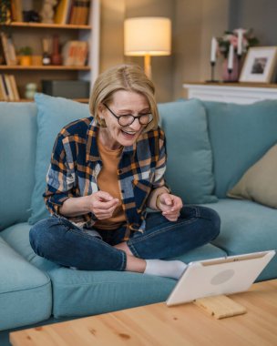 Yetişkin sarışın bir kadın beyaz kadın evdeki kanepeye oturup dijital tablet kullanarak internet üzerinden sohbet ediyor ve uygulamayı gerçek kişilere kopyalıyor.