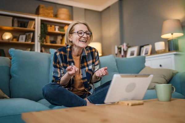一个成熟的金发女人坐在家里的沙发床上 用数字平板电脑在网上聊天聊天 — 图库照片