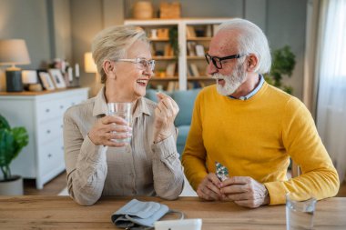Üst düzey çift kadın ilaç ya da vitamin tedavisi alırken kocası beyaz adam evde masada oturuyor. Gerçek insanlar, aile emekliliği ve sağlık konsepti.