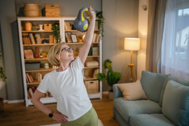 Evde kettlebell Girya Rus çanıyla antrenman yapan olgun bir beyaz kadın. Dairesinde sağlıklı yaşam tarzı kavramında kadın egzersizi yapıyor.