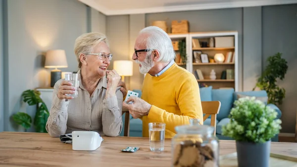 上了年纪的夫妇女人吃药或补充维生素治疗 而她的丈夫老的高加索男人则坐在桌旁真正的人家庭老人退休和健康的概念 — 图库照片