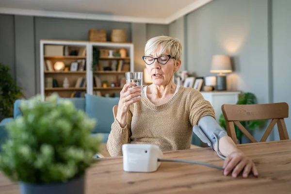 一人の女性シニア原因女性は自宅に座っている間に健康結果を確認するために手に血圧装置を使用して水のガラスを持っています本物の人々の健康管理の概念 — ストック写真
