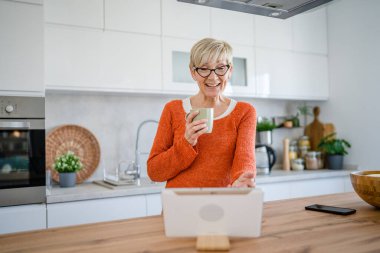 Yetişkin, sarışın, beyaz, son sınıf öğrencisi bir kadın dijital tablet kullanarak internet üzerinden sohbet ediyor.