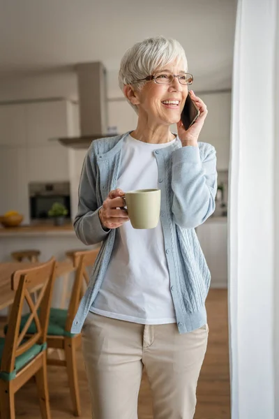 一人の成熟したシニア女性が自宅の窓に立つコールを作るために携帯電話のスマートフォンを使用して実際の人々毎日ルーチンスタンド明るい部屋で短い灰色の髪の摩耗セーターと眼鏡 — ストック写真