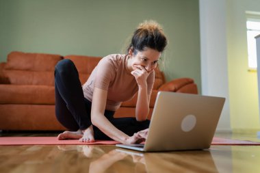 One woman adult caucasian female sitting on the training mat at home using laptop computer to browse internet for online guided training course real people self care concept copy space