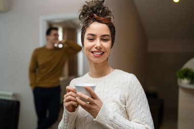 One woman young caucasian female standing in her apartment with cup of coffee while her husband or boyfriend stand in background daily morning evening couple routine real people copy space happy smile