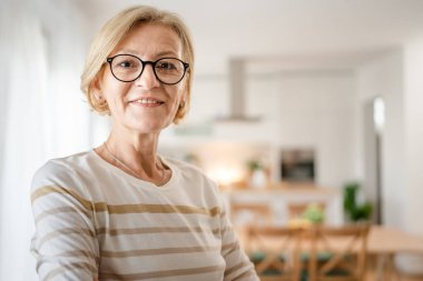 Evde gözlüklü, olgun, beyaz bir kadının portresi mutlu bir gülümsemeyle kameraya bakıyor parlak odada kendine güvenen süveter giyiyor.